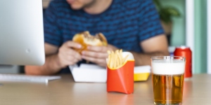 Cropped head of a white male eating burger, fries, soda and beer in front of his computer.
