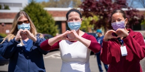 Three women nurses wearing masks and showing a heart sign outside a health facility
